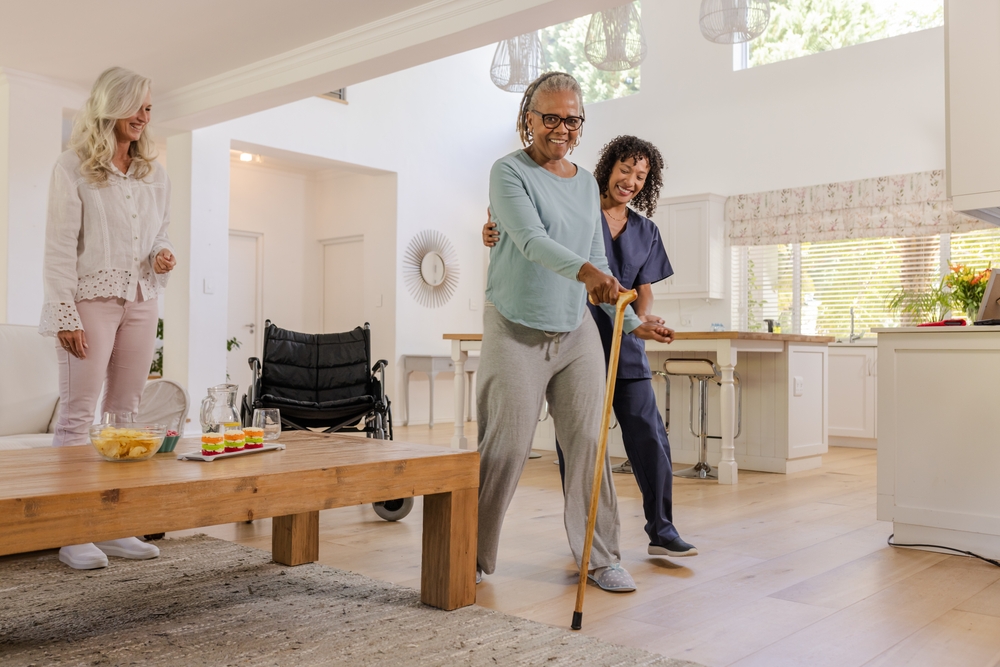 A happy senior woman being helped by a caregiver at home, with a family member in the background.