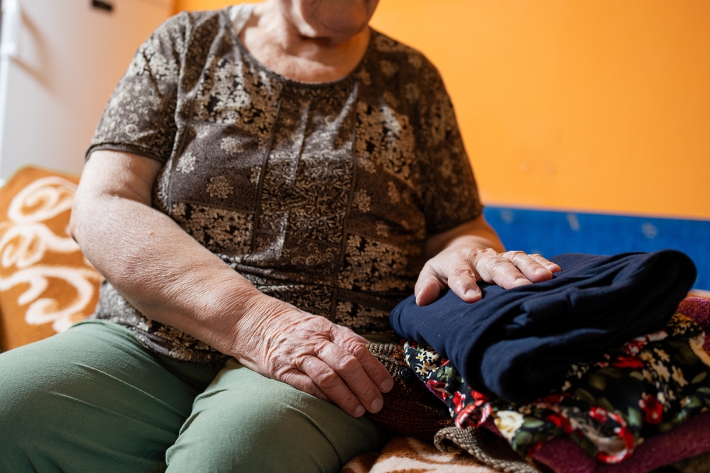 A senior sitting on a bed next to a set of folded laundry.