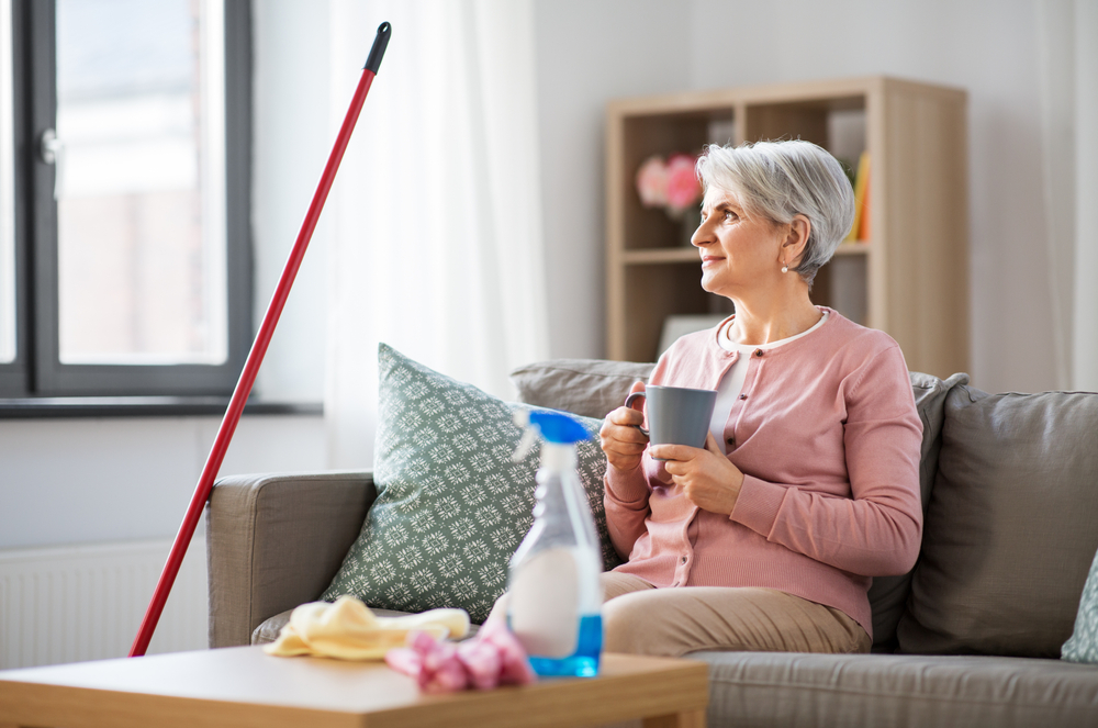 A senior sitting on a couch next to cleaning supplies, drinking a mug of coffee.